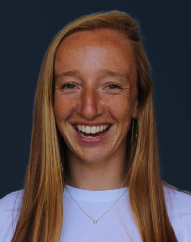 A person with long hair, smiling, wearing a white shirt and a necklace, against a dark background.